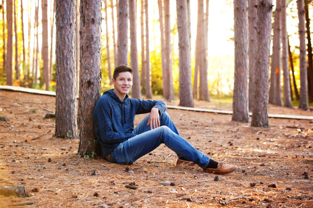 High School Senior Guy sitting by tall pine trees with sun setting in background