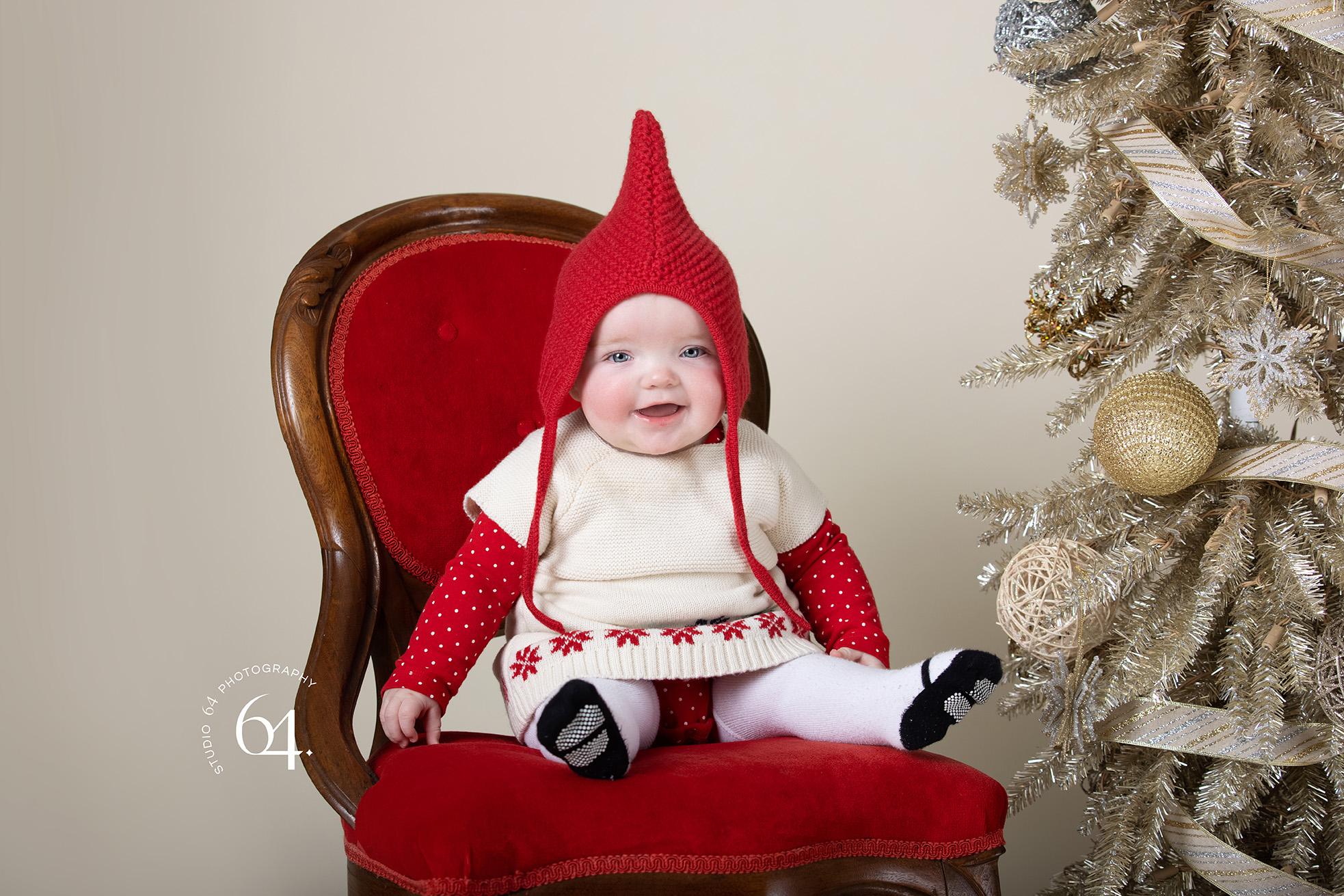 Little Girl sitting on a red chair with a red stocking cap on next to a Christmas Tree