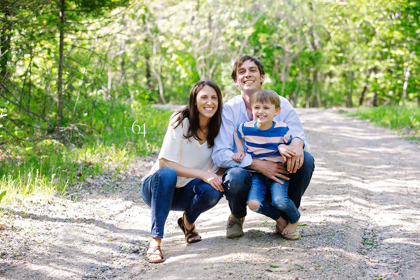 Family of 3 outdoors in the summer time squatting down on a gravel road with big smiles.